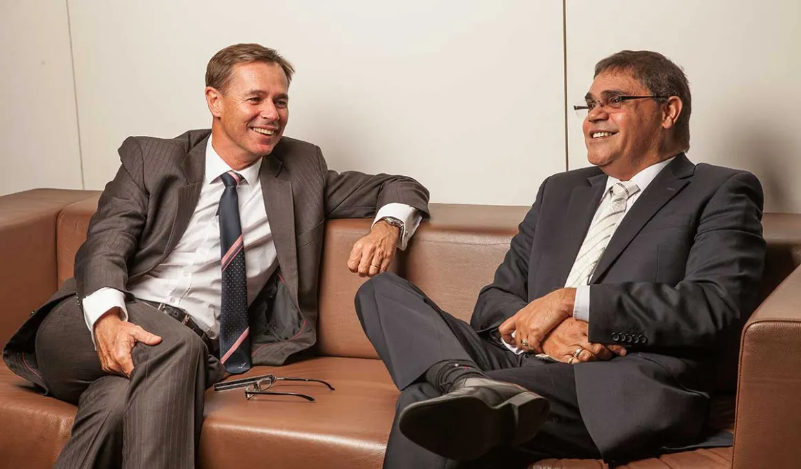 Man on left in brown suit and another man on right in dark grey suit sit on a brown couch with legs crossed and smiling.