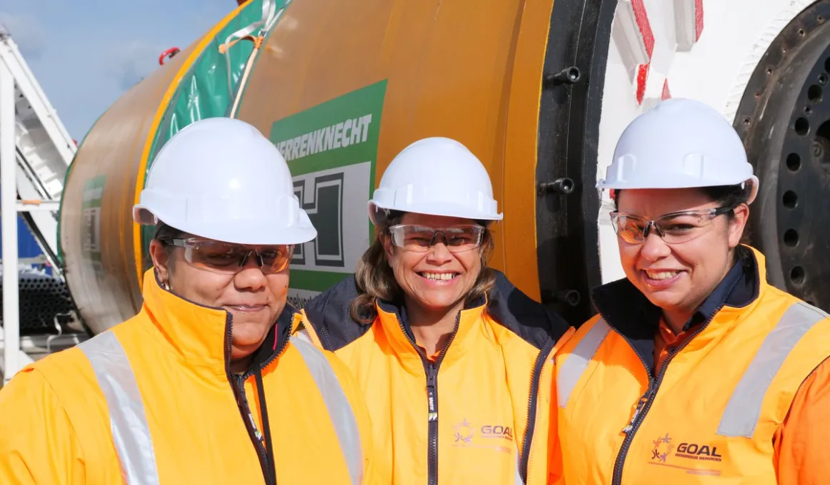 Three smiling Indigenous women in yellow jackets and white helmets stand in front of a large yellow, green and white machine.