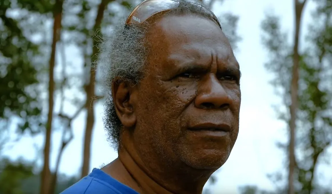 A head photo of an Indigenous man in blue shirt with sunglasses on his head. In the background are trees and sky.