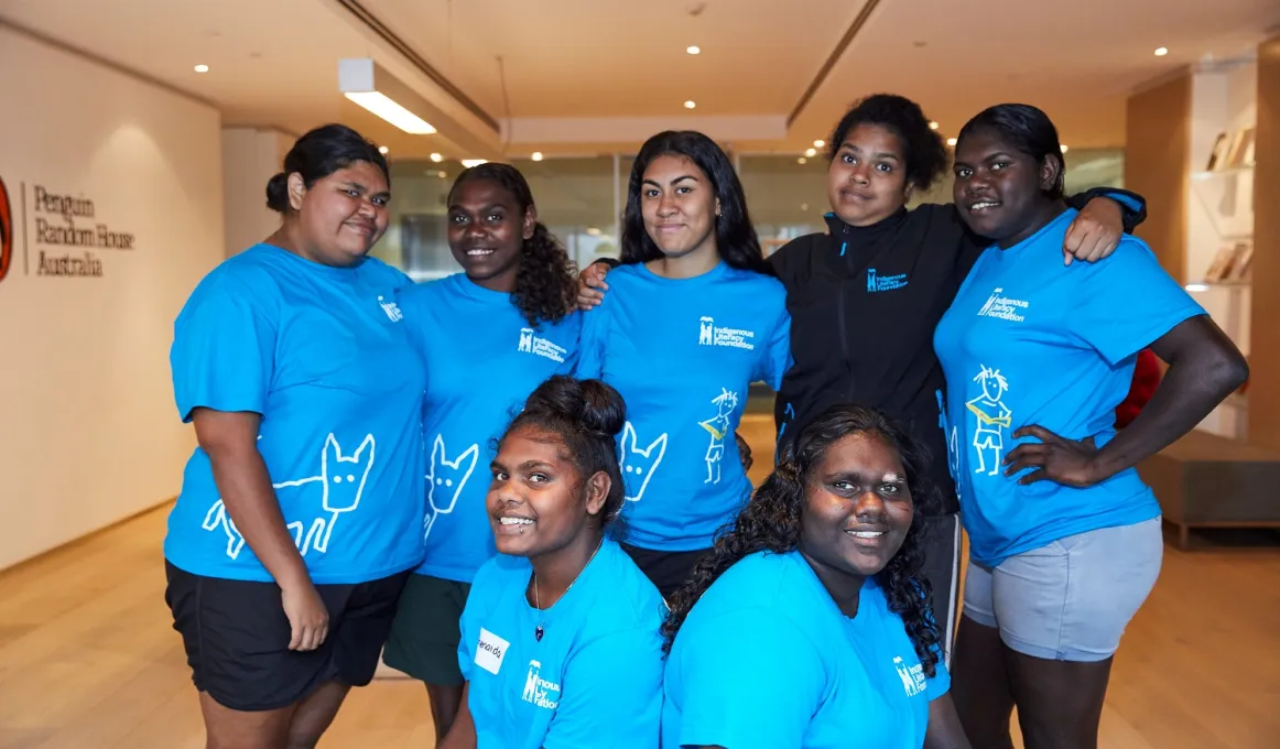 Seven young Aboriginal women, most wearing blue t-shirts stand in a group in a room with a board floor and a book case in the background. At left on the wall are the words Penguin Random House Australia.