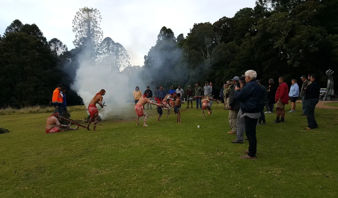 Indigenous dancers in full traditional dress perform next to a fire on a grassed area in front of onlookers. In the background are tall trees.