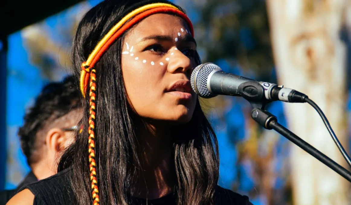 A young Indigenous woman with white dots painted across her face and wearing a black, red and yellow band around her head and long black hair stands in front of a microphone. In the background are trees and blue sky.