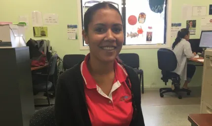 Young Aboriginal woman in red shirt sits in an office with equipment and her colleague in the background. The floor is grey and the walls green in colour.