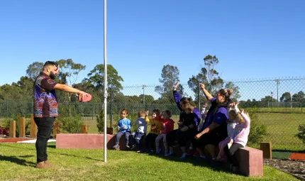 Group of children and adults sit on a low wall. In front is a grassed area on which is a pole. On the other side of the pole stands an Aboriginal man. In the background is a fence and trees and more grass.