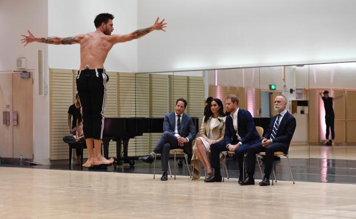 4 people (Duke and Duchess of Sussex and two other men) watching Bangarra Dance Theatre's rehearsal for their up coming tour.In the background a piano and mirrors hung on the wall. 