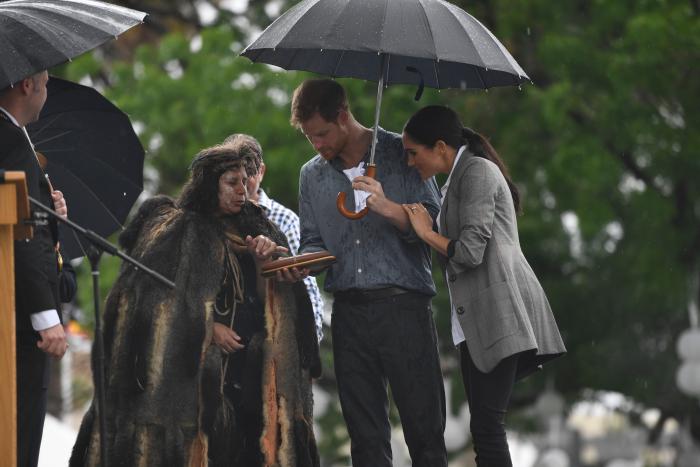 3 people (The Duke and Duchess of Sussex and Aboriginal Woman) viewing tapping sticks in the rain under umbrellas.