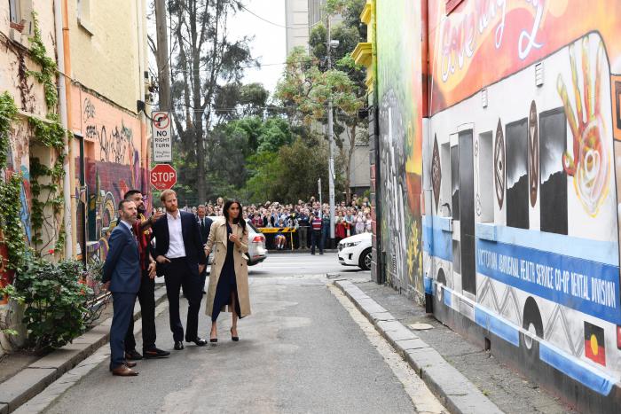 4 people (Duke and Duchess of Sussex, an aboriginal man Robert Young and third man) stand in an alley viewing colourful murals on the walls lining the alley. In the background are cars, a crowd of people, and trees behind the crowd. 