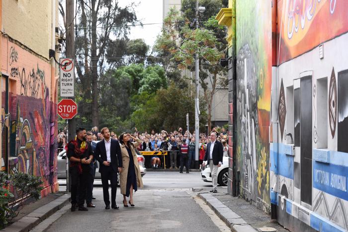 4 people (Duke and Duchess of Sussex, an aboriginal man Robert Young and third man) stand in an alley viewing colourful murals on the walls lining the alley. In the background are cars, a crowd of people, and trees behind the crowd. 