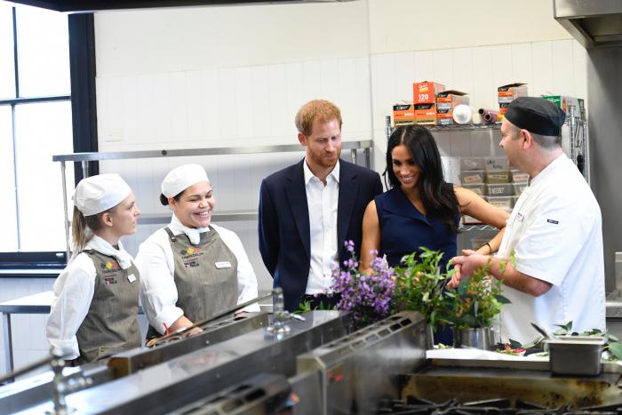 5 people (The Duke and Duchess of Sussex, Executive Chef of restaurant Charcoal Lane and 2 female students) standing in the restaurants kitchen in front of native herbs and plants. 