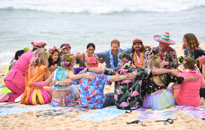 Large group of people including The Duke and Duchess of Sussex in bright colour clothes sitting down in a circle on the beach smiling and hugging. 