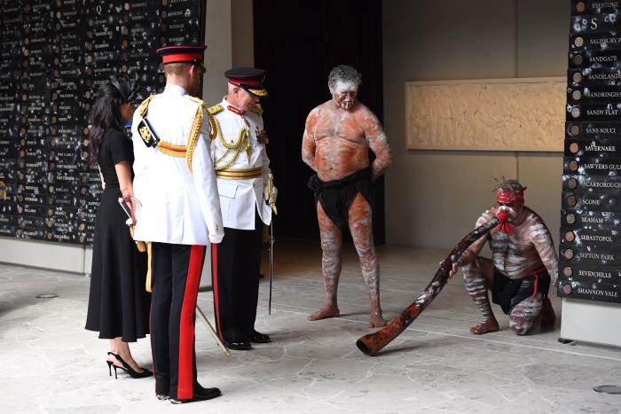 5 people (The Duke and Duchess of Sussex, NSW Governor David Hurley, two Aboriginal men in traditional painting one playing the didgeridoo on the ground at the opening of the ANZAC memorial. 