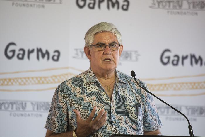 Elderly man with grey hair wearing glasses and an open neck shirt with Indigenous designs stands at a pulpit with micropone. The backdrop is white with the words: Garma, Yothu Yindi Foundation and some long wavy orange lines.