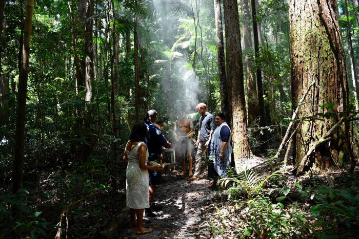 The Duke of Sussex receiving a traditional Welcome to Country Smoking Ceremony from 5 Indigenous men and women standing in a forrest with smoke arising from the ground.