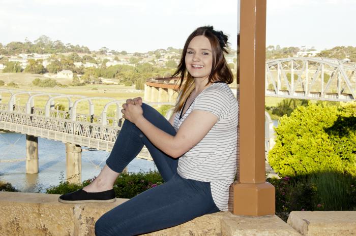 Young woman sits on a wall with her back to a pillar. She is dressed in dark slacks and light shirt. In the background is a tree, a river, two bridges and houses.