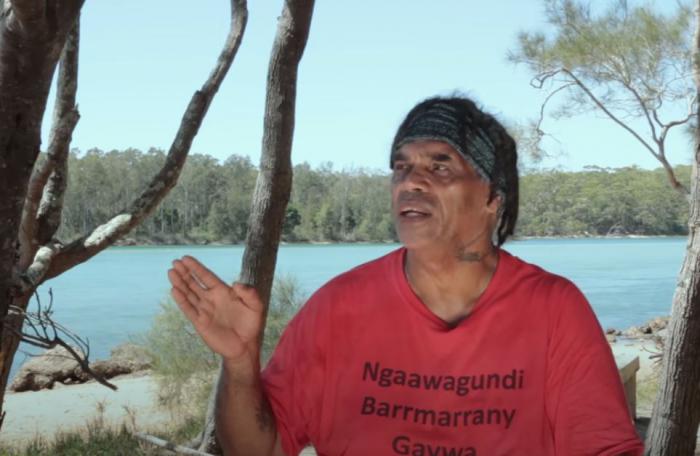 A man wearing a bandana and a red t-shirt looks to the right with his right hand raised. His shirt has the words Ngaawagundi Barrmarrany. In the background are trees, a river and beyond that, a forest of trees. 
