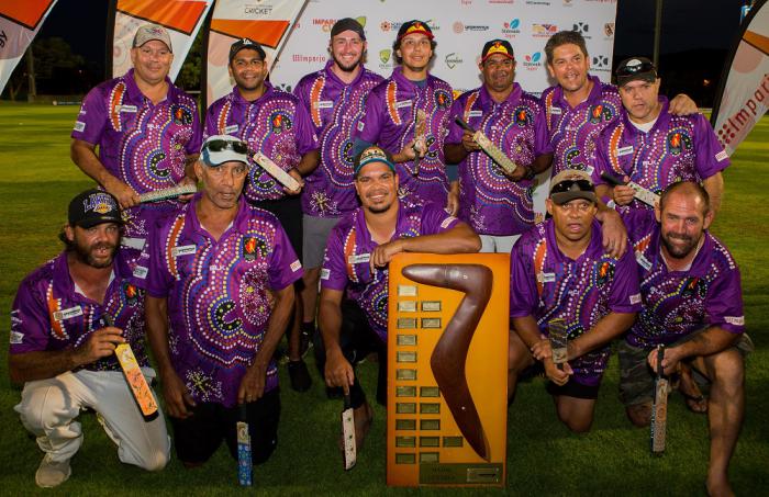 Group of men in purple jerseys featuring Indigenous designs, stand or kneel around a large trophy featuring a boomerang on a large wooden plaque with small metal plates.
