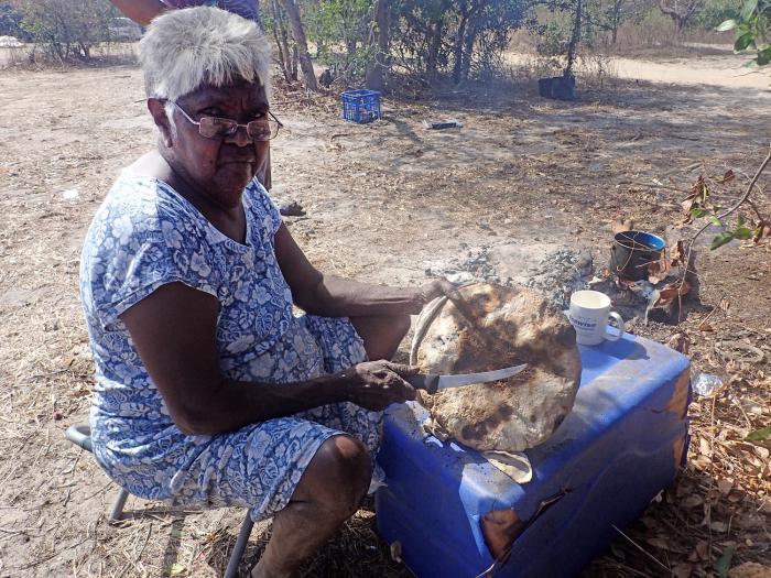 Mature Aboriginal woman in blue and white dress sits at a small table with a knife in one hand and a loaf of damper in the other.