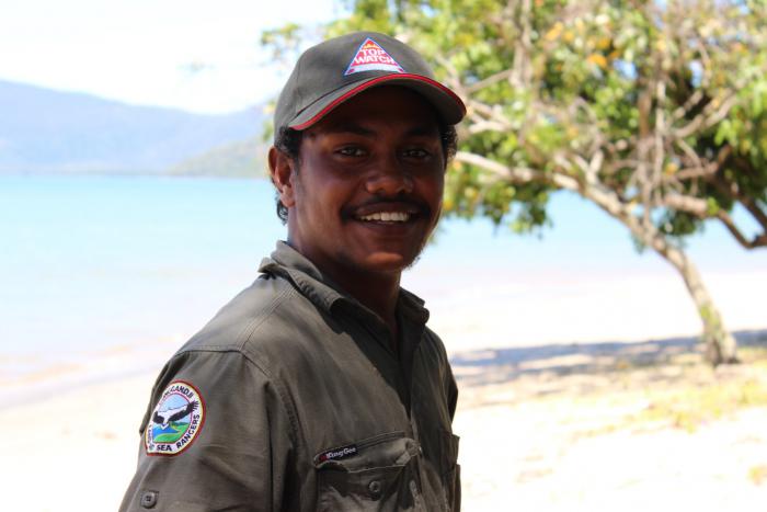 Indigenous young man dressed in ranger clothing standing near tree with body of water and hills in the background.