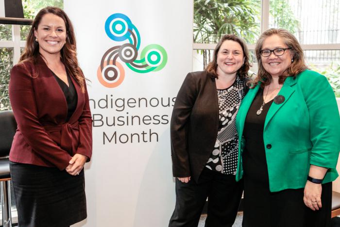 Three women in business wear stand next to a large poster with the words Indigenous Business Month and includes a logo with four circles attached by curved lines. In the background are windows beyond which are trees.