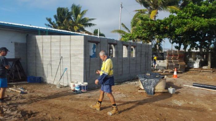 Two men work near newly built grey block addition to white building with trees in background.