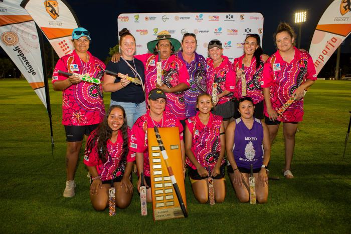 Group of women in pink and purple jerseys featuring Indigenous designs, stand or kneel around a large trophy featuring a digeridoo on a large wooden plaque with small metal plates.