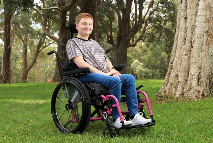 A young woman sits in a wheelchair on grass. In the background are trees.