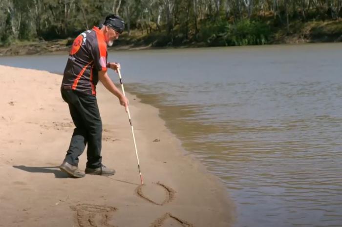 A middle-aged man in dark trousers and red and black shirt stands next to a river and holds a long stick with which he draws lines in the soft sand bank. In the background is more of the river and trees on the opposite bank.