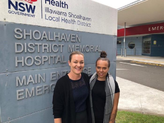 Two women stand in front of a large sign featuring the words Shoalhaven District Memorial Hospital. The woman at left wears a dark cardigan and dress and the one on the right wears a dark dress with a grey and white scarf about her neck.