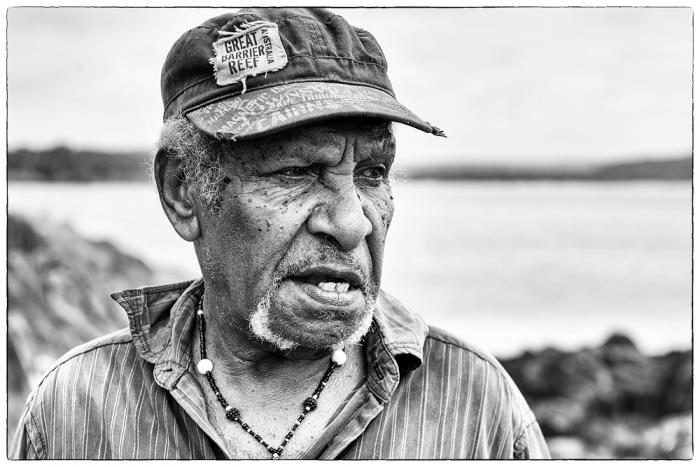 An elderly man in a baseball cap and lined shirt looks to left of camera. In the background are rocks and water.