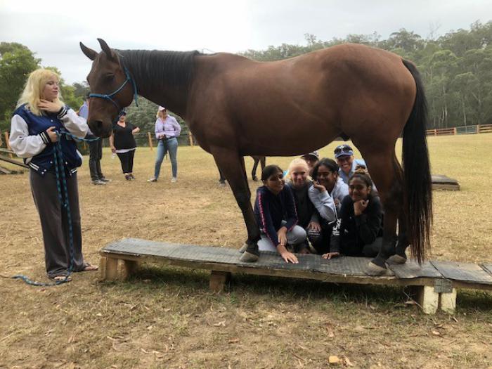 Young girls and two police officers squat behind a horse but are seen through the gap between the front and rear legs. At the head of the horse is a young woman holding its reins. In the background are more people, a grassy area and trees.