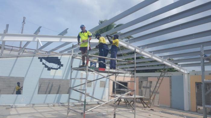 Three men in workwear standing on scaffolding below steel supports which form part of roof construction.