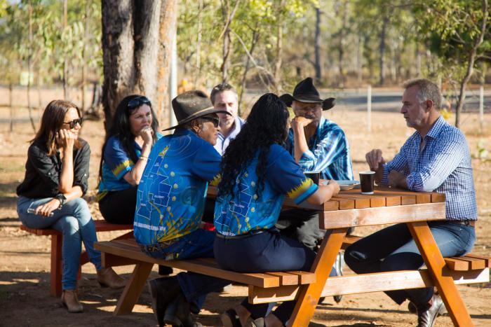 Group of people sit around a picnic table on dry ground. In the background are trees and fence posts.