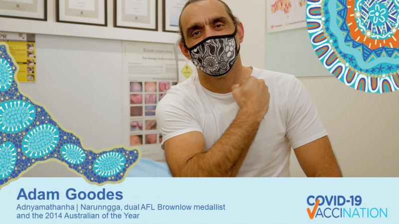 A man sits in a Doctors office. He is wearing a white t-shirt and a black and white patterned face mask. He has one arm held across his chest to show where his upper arm where he was vaccinated