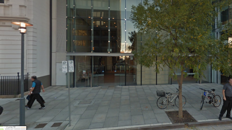 Glass front of building with grey pavers on the footpath and a tree, 2 bikes and two people in front of it.