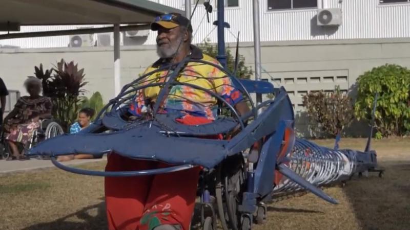 An elderly man sits in a wheel chair that forms part of a long metal contraption in the shape of a hammerhead shark. In the background are two women, grass, shrubbery and a building.