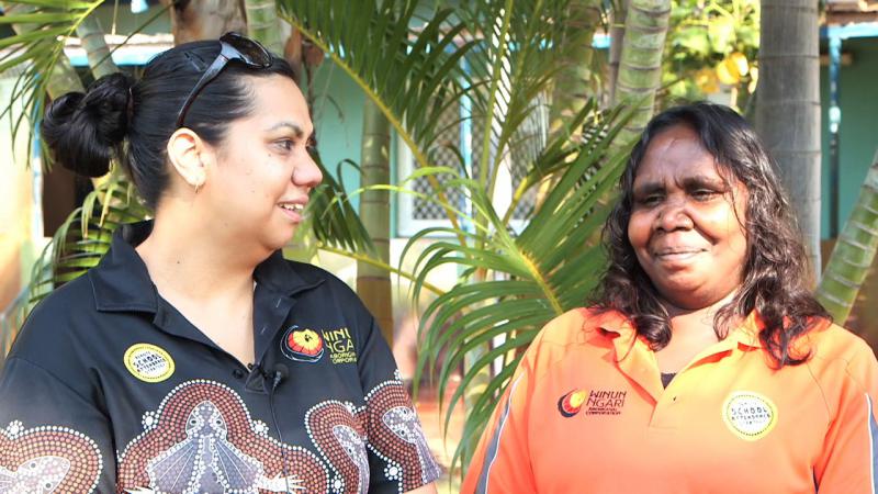 Two Indigenous women standing in front of a palm tree, one wearing a black shirt with Aboriginal design and the other an orange shirt.