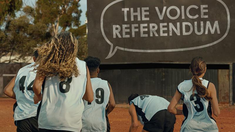 An image of four people wearing sporting jerseys running towards a sign that reads 'The Voice Referendum'. 
