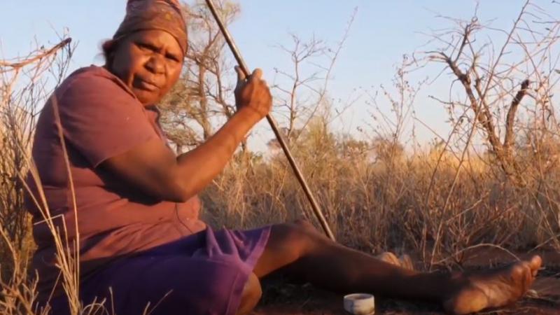 A mature Aboriginal woman dressed in top and skirt sits on red soil in amongst long grass. She holds a metal rod in her right hand.