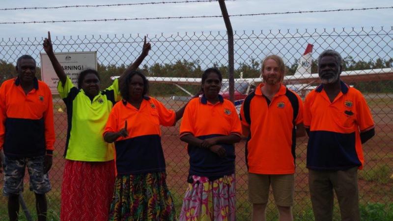 Five Aboriginal people and one non-Aboriginal person are standing in front of a remote airfield. In the background is a small airplane.