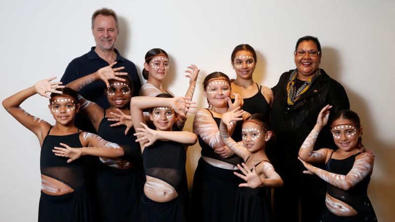 Non-Indigenous man and Aboriginal woman stand in front of white background with young female Indigenous dancers between and in front of them. All are dressed in black and the dancers wear white face paint.