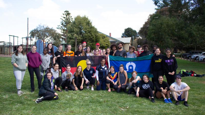 Group of Indigenous youth sit or stand on grass, some holding the Aboriginal flag and Torres Strait Islander flag. In the background is a building, cars and trees.