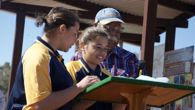Two young Aboriginal girls dressed in blue and yellow polo shirts stand at a podium with an elderly Aboriginal man in blue cap and blue and red shirt. In the background is a small metal shelter.