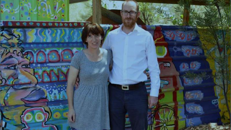 A woman in a grey dress and colourful tights and a man in white shirt and blue trousers stand in front of a painted fence featuring a face, a bicycle and other designs of blue, green, red, yellow and purple.