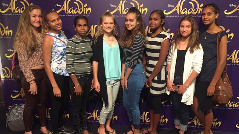 Eight young Indigenous women standing on a carpeted floor in front of a poster wall with multiple displays of the word ‘Aladdin’.