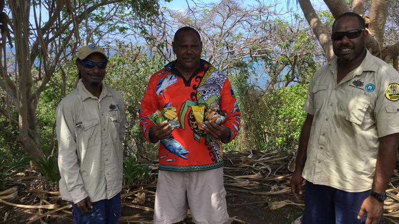 Three Indigenous men, two in ranger shirts, stand on bare soil in amongst trees. The man in the middle holds some seed packets. In the background is the ocean. 