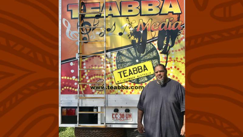 Aboriginal man in grey shirt stands in front of the rear door of a truck. The door is painted with red, yellow and black designs and includes the following words: TEABBA Media, TEABBA N.T. Outback Australia, www.teabba.com