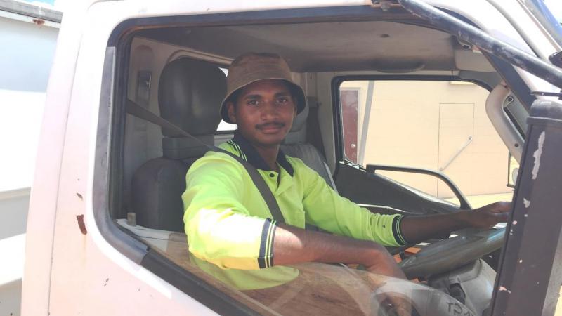 An Indigenous young man wearing work wear sits in the cab of a truck.