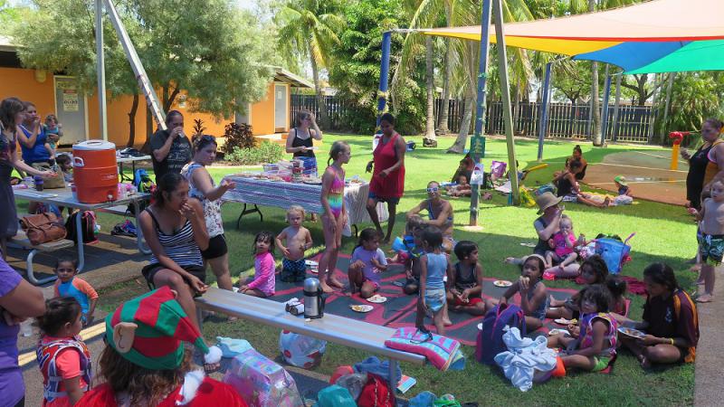 Large group of children and adults sit on grass and at tables under shade, dressed in swimming attire and other causal clothes. In the background are trees, shade covers and a building.