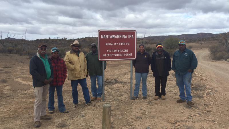 Seven Aboriginal men and women dressed in warm clothing stand next to a red sign just off a dirt road. In the background are bushes, trees and hills. The sign says Nantawarrina IPA – Australia’s first IPA – Visitors welcome – No entry without permit.