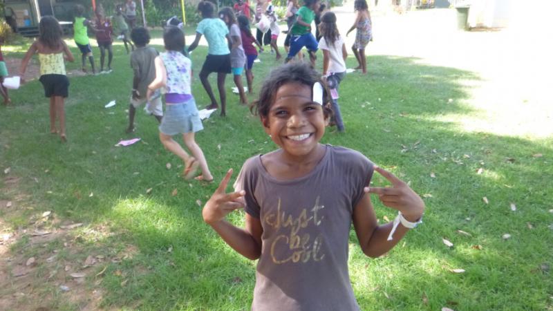 Young Aboriginal girl smiling at camera and other children in background. All are playing on the grass.